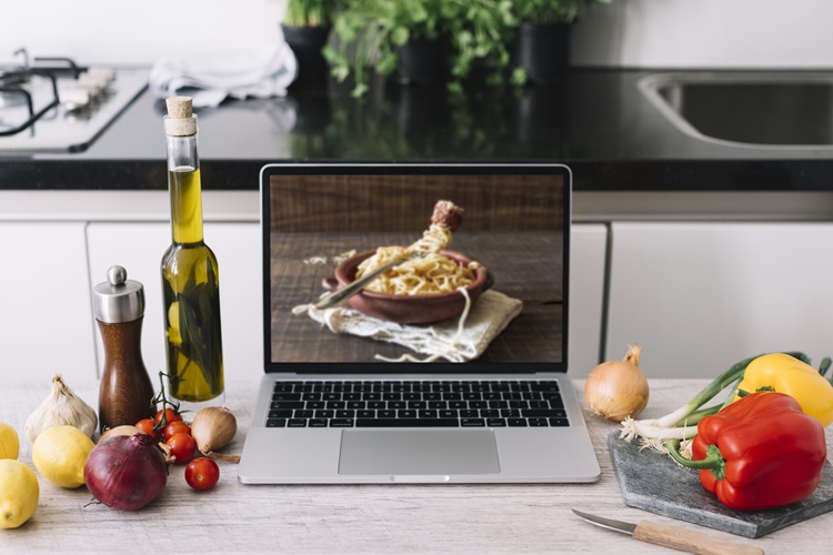 Vegetables and condiments on kitchen worktop scattered around a laptop which shows an image of a bowl of pasta 